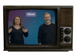 Ein Fernseher, wo man zwei Personen sieht: Anke (links) und Miki (rechts). Sie lächeln.